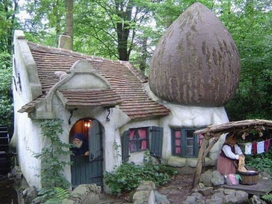 A white & brown hobbit house, in front of which a woman washes and hangs linens to dry. Trees surround it all.
